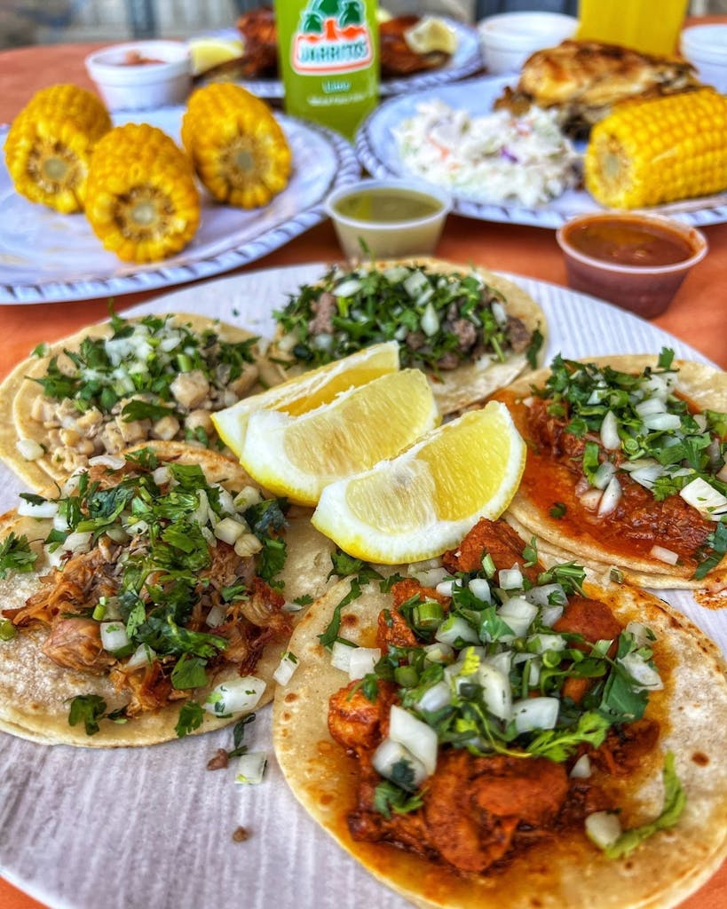 Close-up of five street tacos with cilantro and onion, lemon wedges, and cups of green and red salsa; corn, coleslaw, and chicken in the background.