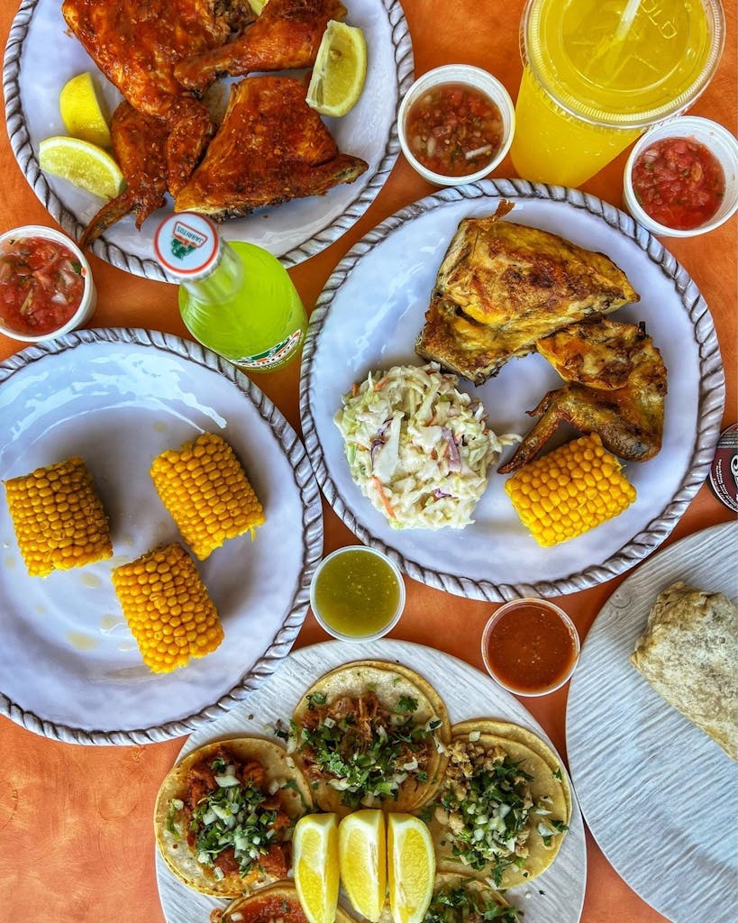 Spread of grilled chicken with lemon, corn on the cob, tacos, coleslaw, salsas, Jarritos, and a yellow drink.