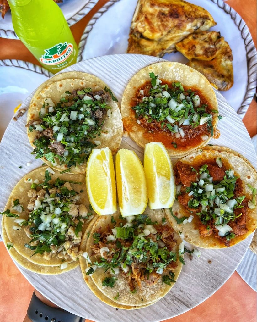 Plate of six street tacos with cilantro and onion and lemon wedges; Jarritos lime soda and grilled chicken in the background.