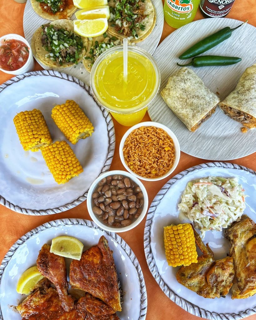 Table spread with tacos, burrito halves and serrano peppers, grilled chicken, corn, rice, beans, coleslaw, lemon wedges, and drinks.