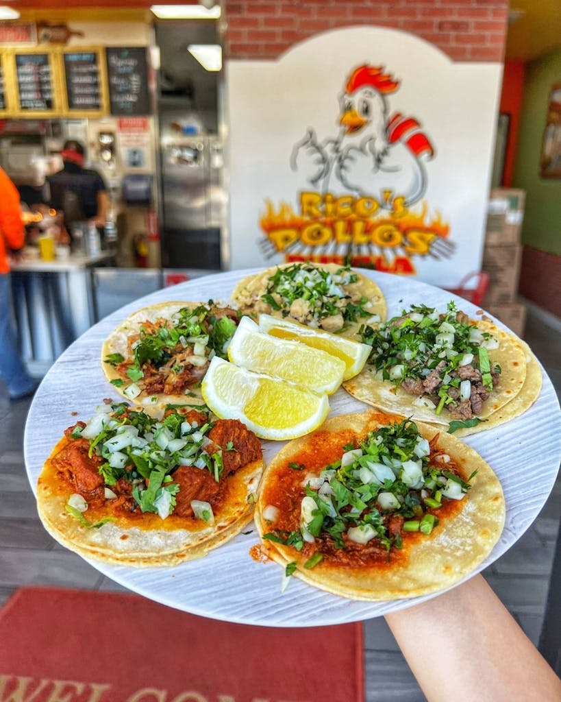Plate of five street tacos with lemon wedges held inside the restaurant in front of the Rico’s Pollos Puebla sign.