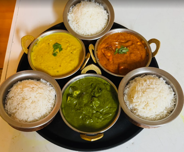 a spread of Indian cuisine, featuring a variety of traditional curries served in brass-handled bowls alongside bowls of steamed Basmati rice