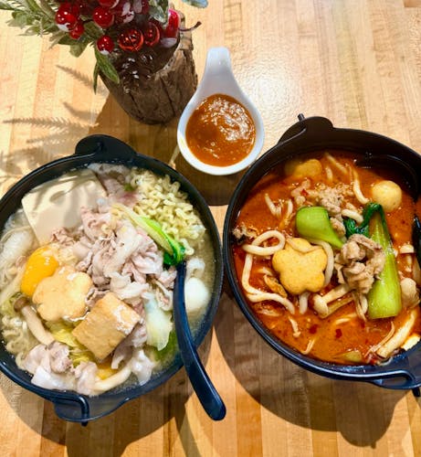 Two noodle hot pots—one clear, one spicy red—topped with pork, tofu, egg, bok choy and mushrooms, with dipping sauce between