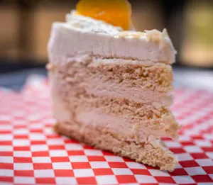 Slice of vanilla layer cake with whipped frosting and a citrus garnish on red-and-white check paper